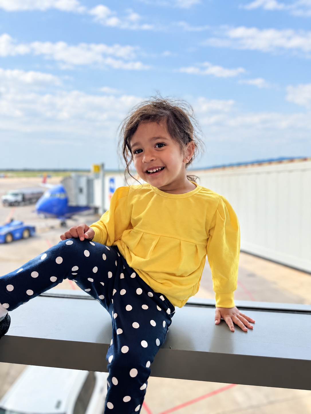 Aria grinning while waiting at the airport, plane outside the window