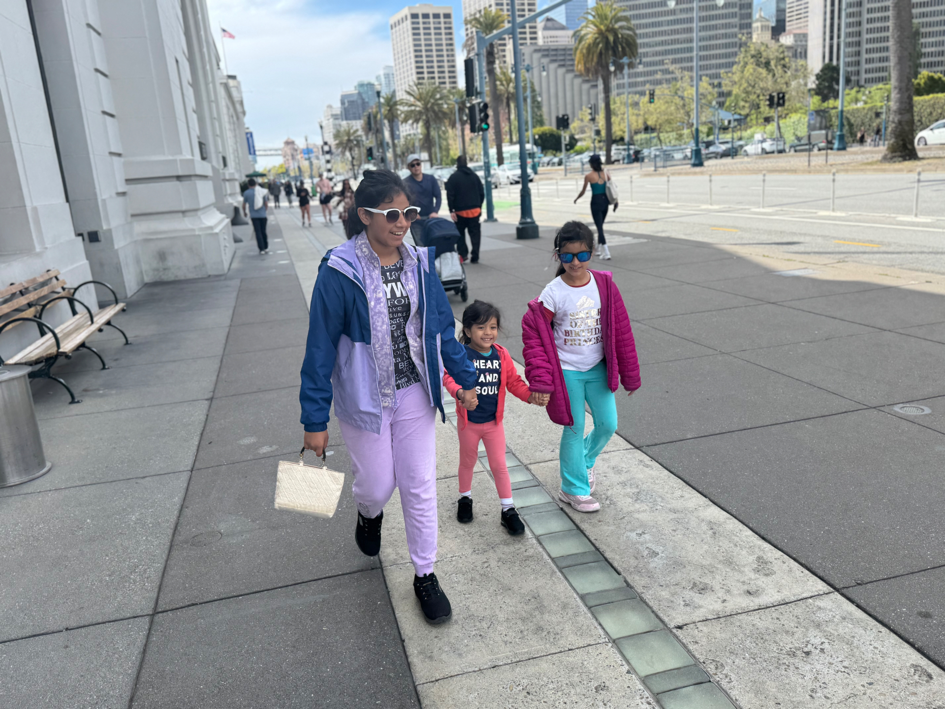 Aria walking hand in hand between her two sisters on a city sidewalk